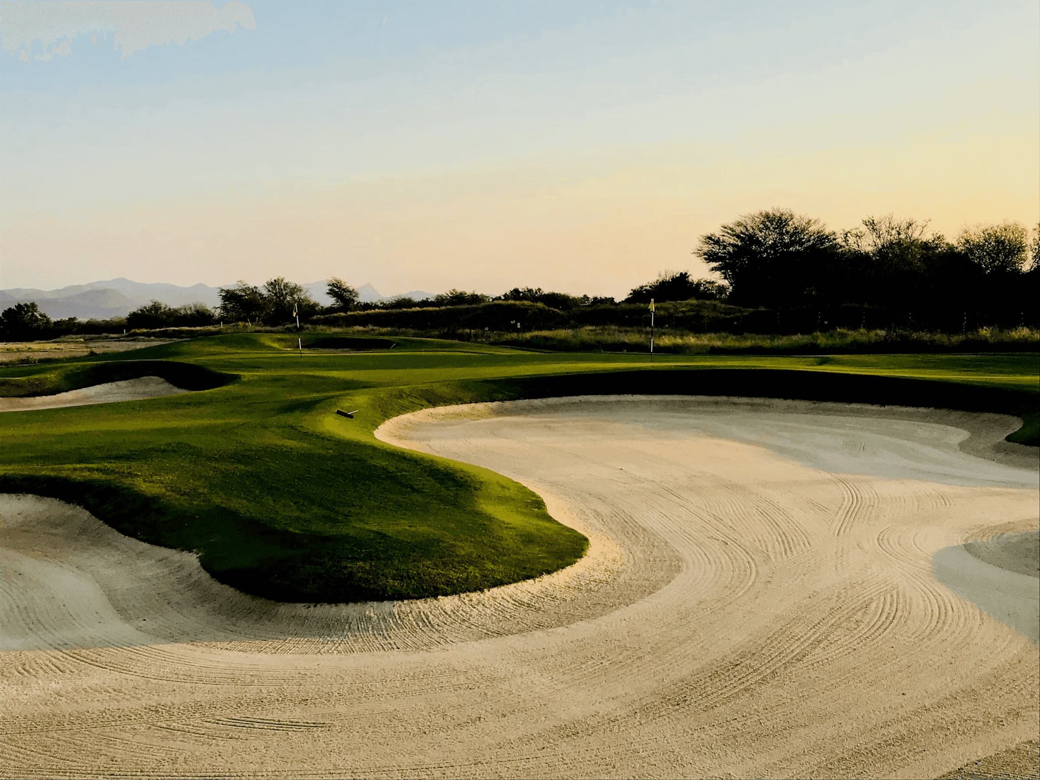A golf course featuring sand bunkers, green fairways, and scattered trees under a hazy sky at sunset.